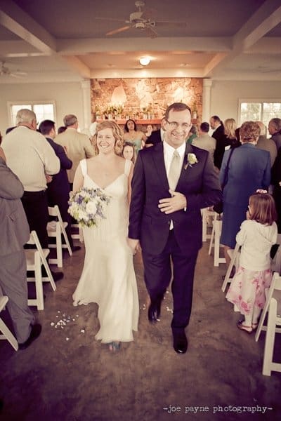 A bride and groom smile while walking down the aisle in a wedding venue. The bride wears a white dress and holds a bouquet, and the groom is in a suit. Guests are standing and facing them, creating a joyful atmosphere.