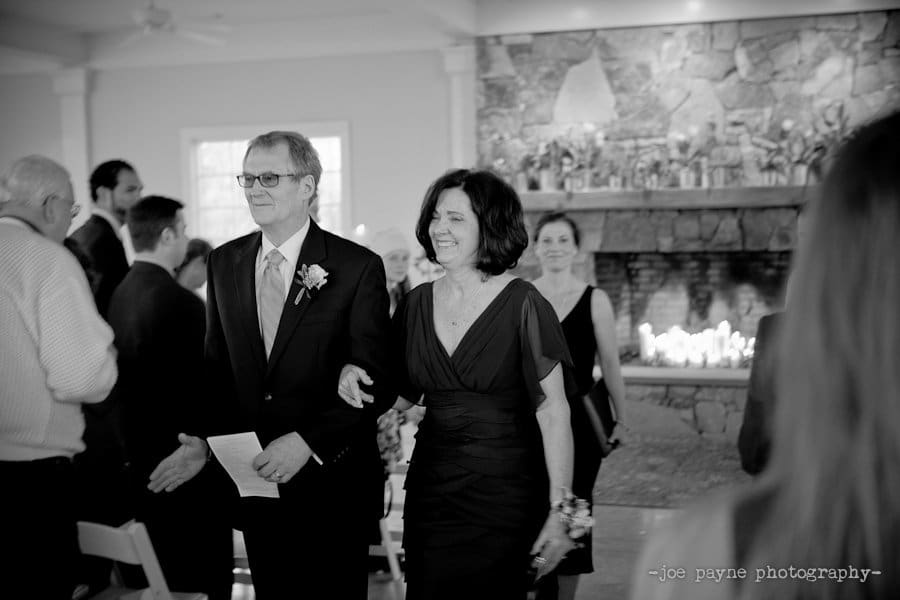 A man and woman walk arm in arm down an aisle at a wedding ceremony. They are dressed formally, with flowers pinned to their clothing. Guests are seated on either side, and a stone fireplace is visible in the background. The scene is in black and white.