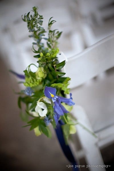 A close-up of a wedding chair decorated with a floral arrangement featuring blue and white flowers, green foliage, and a dark blue ribbon, set against a blurred background.
