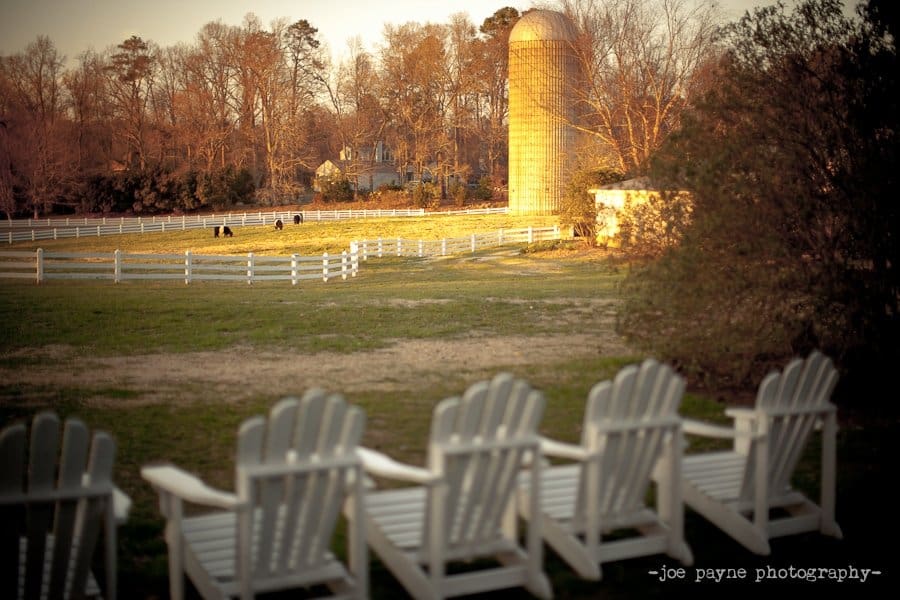 A serene farm scene at sunset with white Adirondack chairs in the foreground. A golden silo and a white-fenced field are visible in the background. Trees in the distance are gently lit by the warm light.