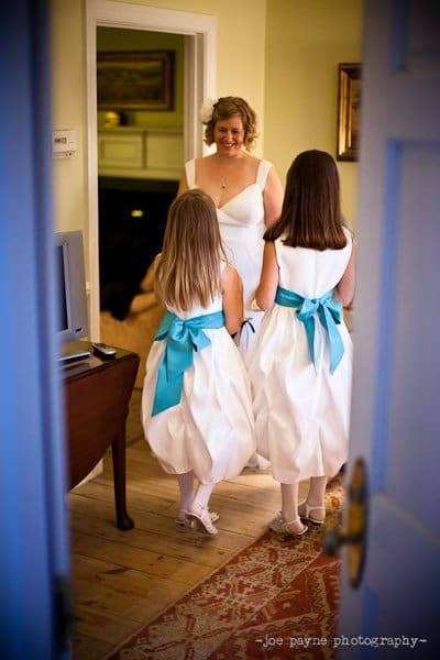 A bride in a white dress smiles at two young girls, likely flower girls, also in white dresses with teal sashes. They stand on a patterned rug in a warmly lit room with a partially open blue door in the foreground.