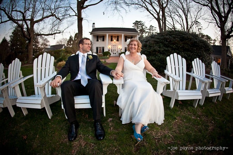 A couple in wedding attire sits joyfully on white Adirondack chairs, holding hands. They are outdoors with a large house in the background. The scene is surrounded by trees under a clear sky.