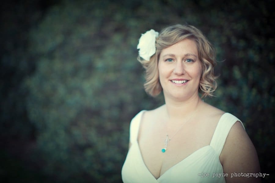A woman with short, wavy blonde hair and a flower in her hair smiles at the camera. She is wearing a white dress and a necklace with a blue pendant. The background is blurred greenery.