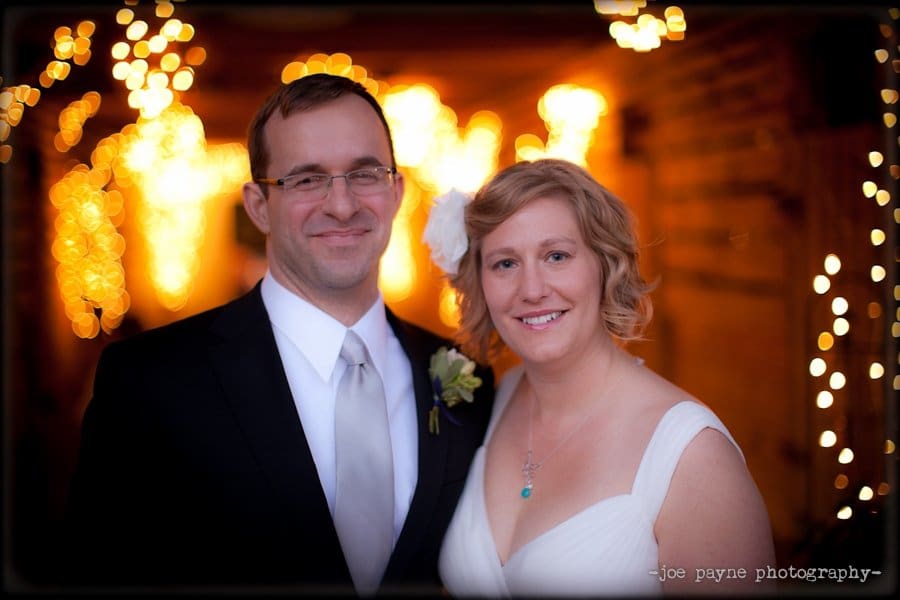 A smiling couple poses in wedding attire. The groom wears a black suit and tie, and the bride wears a white dress with a floral hair accessory. Warm, glowing lights form a bokeh effect in the background, creating a romantic atmosphere.