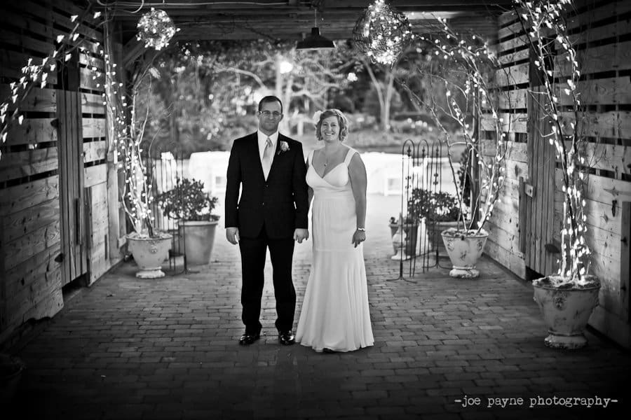 A couple stands under a wooden archway adorned with string lights and potted plants. The woman is in a white dress and the man in a suit, both facing forward. The scene has a romantic, rustic ambiance.