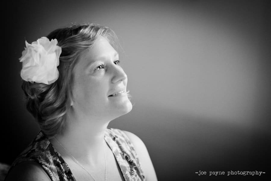 Black and white portrait of a smiling woman with a large flower in her hair, looking upward. Soft lighting highlights her features, with a blurred background. Joe Payne Photography watermark in the corner.