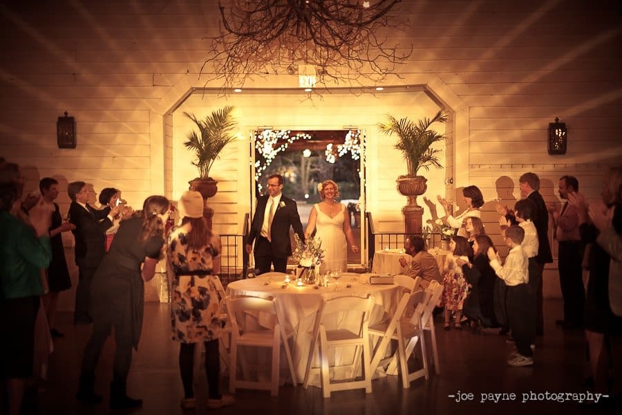 A couple enters a warmly lit reception hall, greeted by clapping guests. The venue is decorated with string lights and plants, creating a cozy atmosphere. Tables are set with white chairs, and the couple stands under a rustic light fixture.