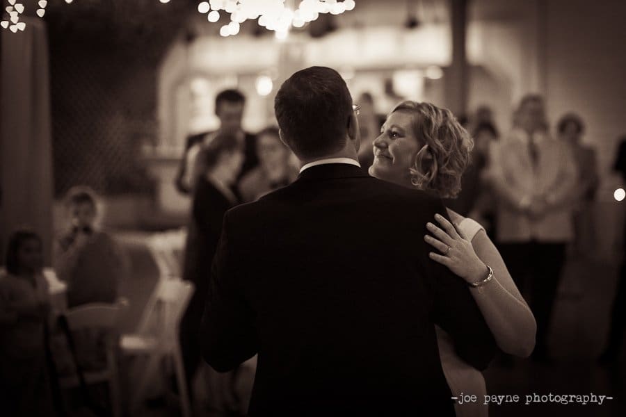 A couple shares a romantic dance at a wedding reception. The setting is warmly lit with blurred guests and lights in the background. The bride smiles, looking over the groom’s shoulder.