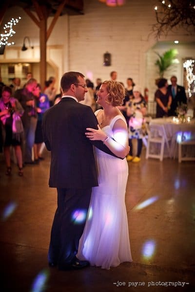 A bride and groom are dancing together in a warmly lit reception hall with guests in the background. The bride is wearing a white dress and the groom is in a suit. Soft, colorful lights dot the floor.