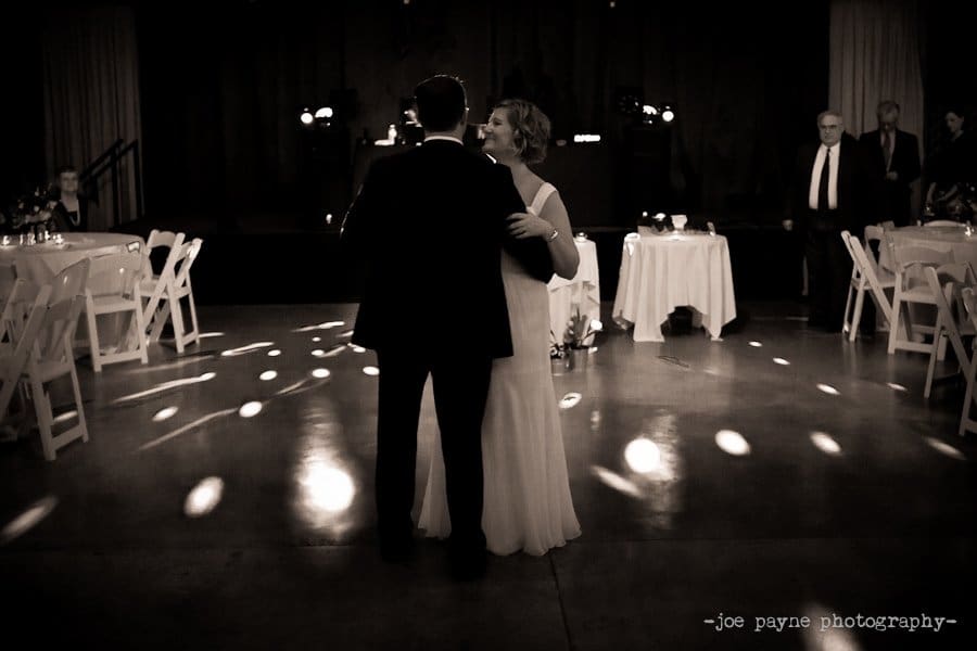 A couple is dancing on a dimly lit dance floor, surrounded by scattered white chairs and tables. The scene has a warm, romantic atmosphere with soft spotlights illuminating the couple.