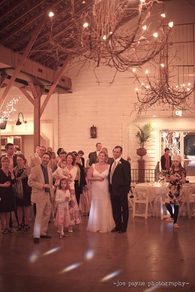A wedding reception in a warmly lit rustic hall. Guests gather around a couple standing together, the bride in a white gown and the groom in a suit. Above, branches and fairy lights hang from the ceiling, creating a cozy ambiance.