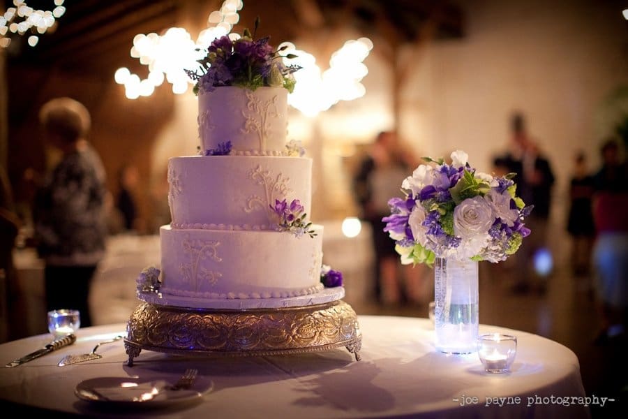 A three-tier white wedding cake with purple floral decorations sits on a table with a silver stand. A matching floral bouquet in a vase and small candles surround it. The background is softly lit, showing blurred guests in a festive setting.