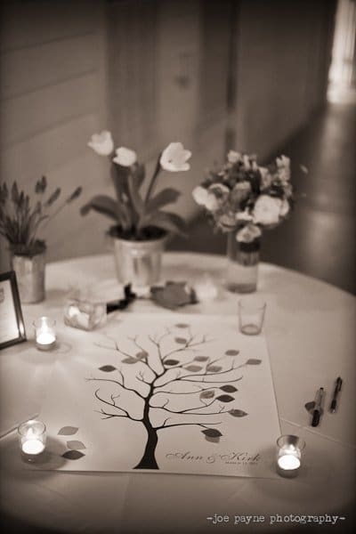 A sepia-toned photo of a round table with a tree-themed guestbook, three small candles, and flower arrangements. Pens are scattered on the table for signing. Soft lighting creates a warm ambiance.