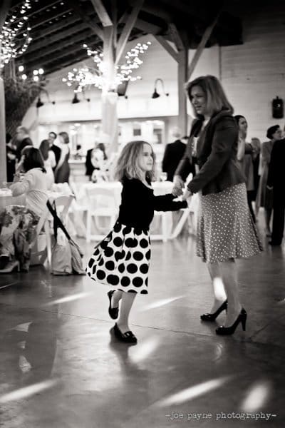 A black and white photo of a woman and a young girl dancing together at an indoor event. The girl is wearing a polka dot dress, and the background shows other people seated at tables and standing under string lights.