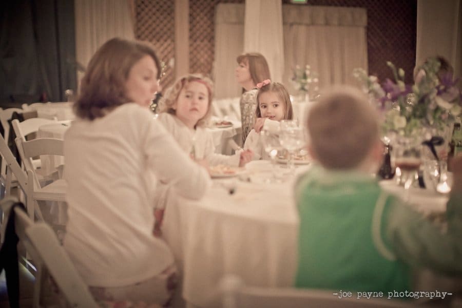 A group of children sit at a round table during what appears to be an indoor event. They are engaged in conversation, and the setting is softly lit with decorative flowers visible in the background.