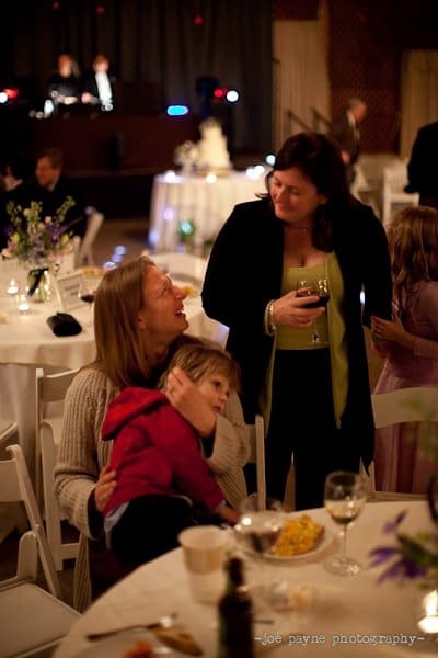 A woman seated at a round table is holding a young child. Another woman, standing nearby, is holding a wine glass and engaging in conversation. The scene is set in a warmly lit event space with decorated tables and people in the background.