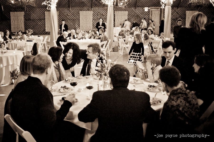 A group of people sit around a table at a formal event. They are engaged in conversation, with plates of food and glasses of wine in front of them. The background shows more guests seated at tables under decorative lighting.
