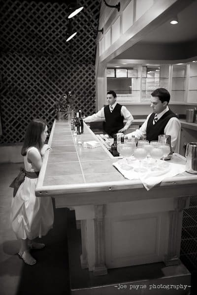 A young girl in a white dress stands at a bar, facing two bartenders dressed in vests and ties. The setting appears to be an event venue with a tiled bar and decorative lattice wall. Black and white photo.