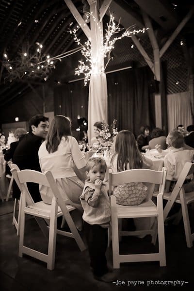 A toddler stands next to a round table with seated guests at a wedding reception in a dimly lit venue. A decorative tree with lights is in the background, adding to the warm ambiance of the gathering.