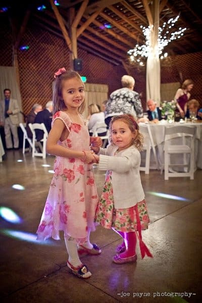 Two young girls in floral dresses hold hands and dance at a wedding reception. The venue has glowing string lights, and guests are seated at tables in the background.