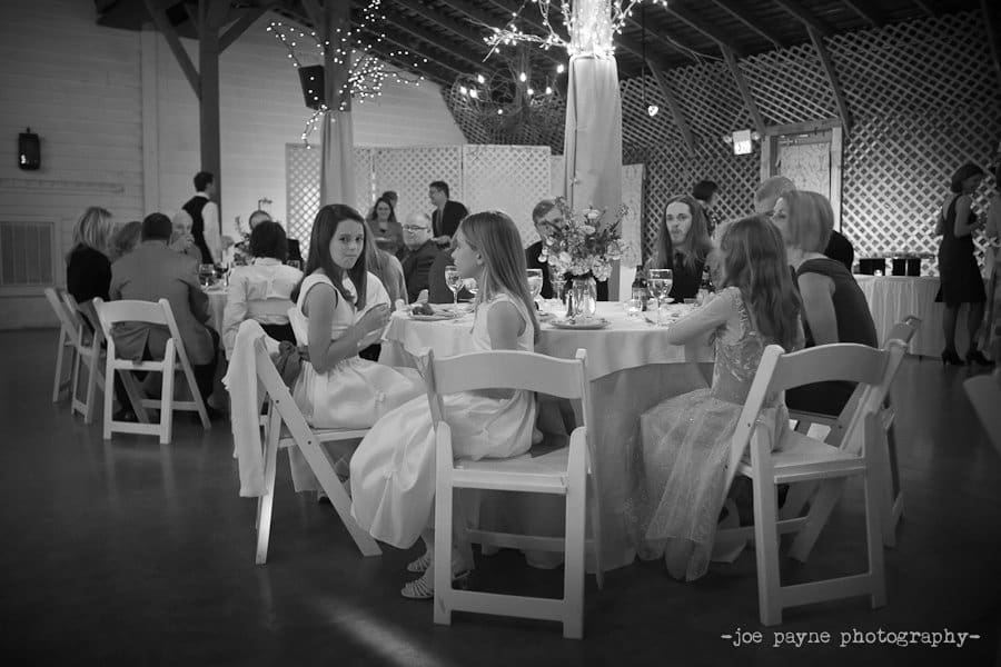 A black and white photo of a festive gathering in a decorated hall. Groups of people, including children in formal attire, are seated at round tables. The space is lit with string lights wrapped around columns, creating a cozy atmosphere.