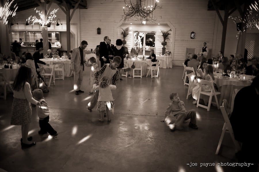 A sepia-toned image of a wedding reception in a hall. Guests, including children, are dancing and interacting in the center. Tables with seated guests surround the dance floor, and twinkling lights decorate the space.