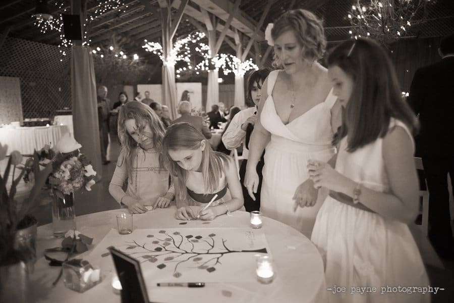 A woman and three children engage in an art activity at a table. The children are drawing on a paper with a tree design. The room is dimly lit, decorated with string lights, and other guests are visible in the background.