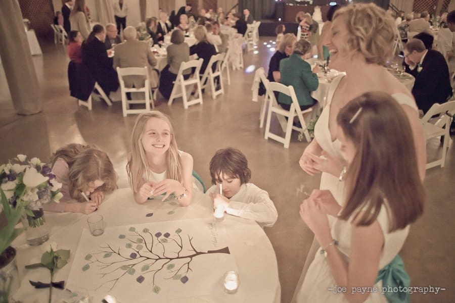 Children gathered around a table at a wedding reception, coloring a tree poster with leaves. Guests are seated at tables in the background, creating a warm and joyful atmosphere.
