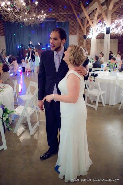 A couple stands together at a wedding reception. The woman, in a white dress, has her arm around the man in a dark suit. Guests are seated at round tables decorated with flowers. String lights illuminate the wooden ceiling above.