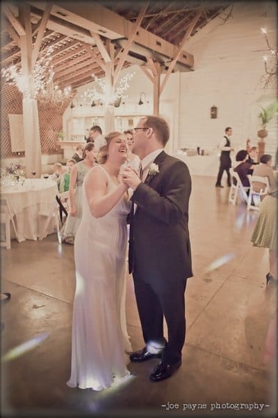 A bride and groom share a joyful dance at their wedding reception. They are smiling and holding hands on a wooden floor under a rustic ceiling with string lights. Guests are seated at round tables in the background.