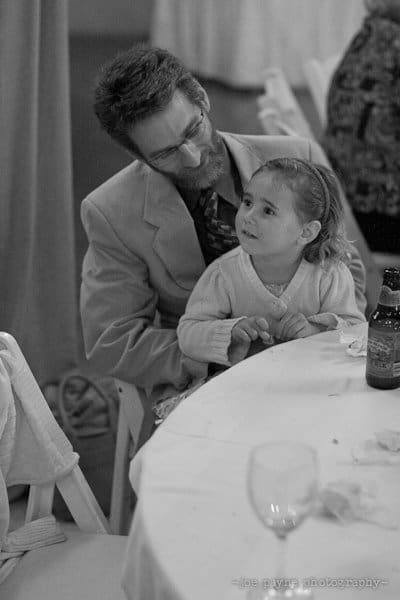 A man with a beard and glasses sits at a table, looking at a young girl beside him. The girl has her hair tied back and is also looking at him. There are empty glasses and a beer bottle on the table. The image is in black and white.