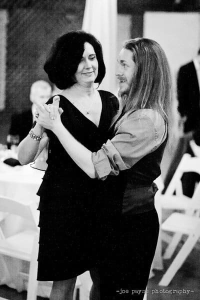 A black and white photo of a man and a woman dancing together at an event. The woman is wearing a sleeveless dress and the man has long hair and is wearing a long-sleeve shirt. They appear to be smiling as they dance.
