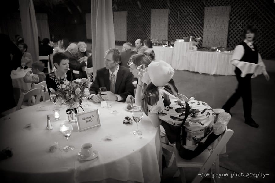 A black and white photo shows a group of people seated at a round table with a Reserved for Family sign. A child playfully leans over to hug one of the seated individuals. In the background, a person walks by carrying a tray.