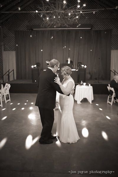 A couple dances together in an elegant, dimly lit venue. They are centered under a chandelier with scattered light patterns on the floor. A DJ booth is visible in the background. The setting appears intimate and romantic.