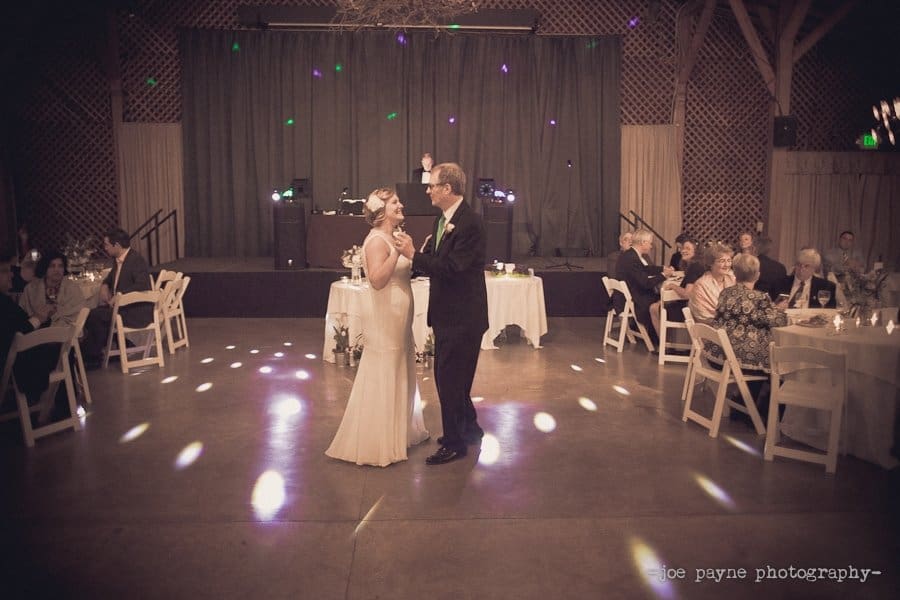 A bride and groom share a first dance in a warmly lit wedding venue. Guests are seated at tables decorated with white tablecloths and candles, observing the couple with smiles. The DJ booth is set up on a stage behind them.