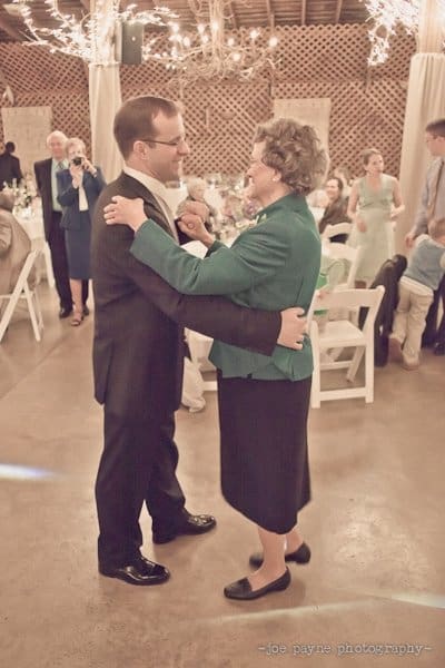 An elderly woman and a man dance together at a wedding reception. They are smiling at each other, surrounded by guests seated at tables. The venue is decorated with string lights.