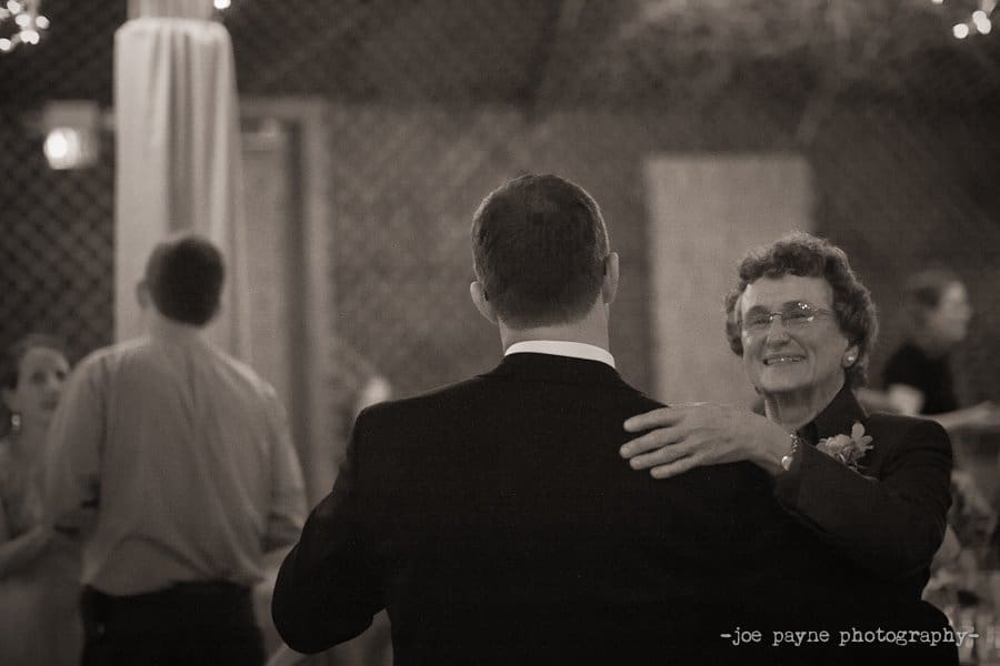 A black and white photo of an elderly woman smiling warmly as she dances with a man in a suit. They are in a softly lit setting with people and string lights in the background.