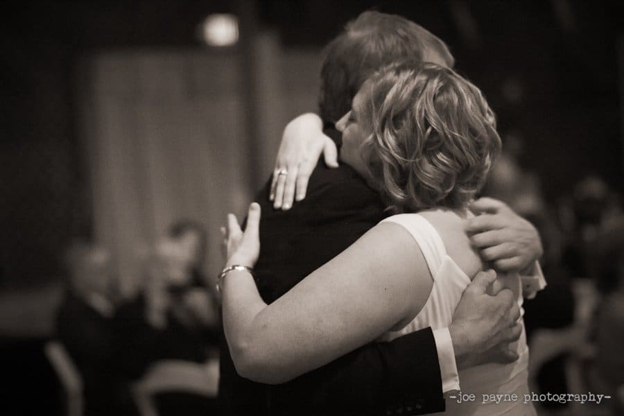 A couple is embracing while dancing in a warmly lit room. The womans back is visible, showcasing her sleeveless dress and short, wavy hair. The man is in a suit. The background is softly blurred, with people seated at tables.