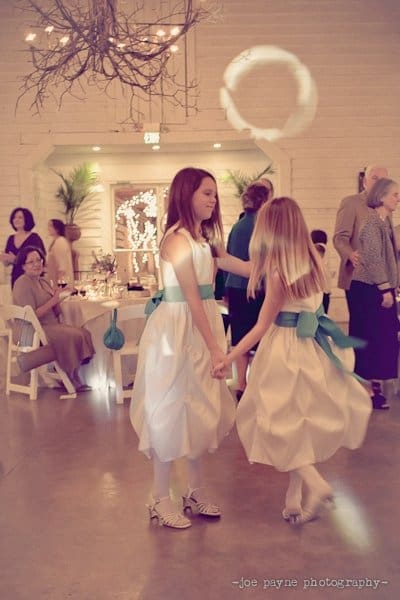 Two young girls in white dresses with blue sashes dance together at an indoor event. Nearby, guests sit at round tables under a chandelier. The room is softly lit, with white walls and a cozy ambiance.