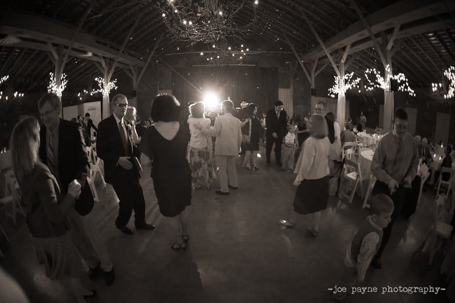 A sepia-toned photo of people dancing at a wedding reception. Guests, dressed in formal attire, move around the dance floor under twinkling lights in a barn-like venue. A bright light shines in the background, creating a festive atmosphere.
