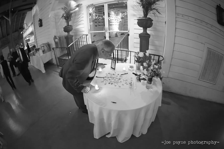 A man leans over a round table with a guestbook and candles, set in a white-paneled room. There are flowers on the table and another group of people in the background.