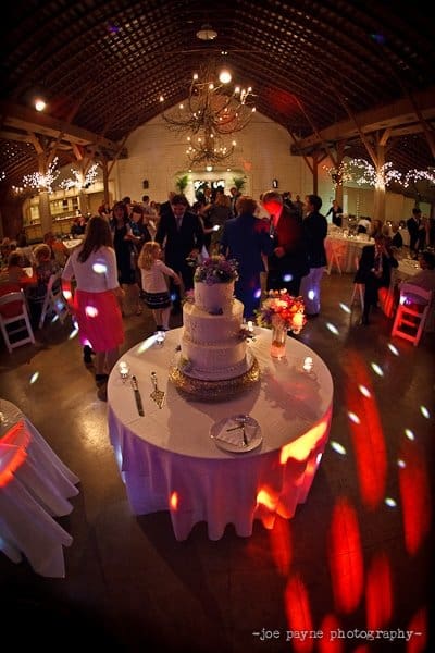 A wedding reception in a barn-style venue with a high ceiling and chandeliers. Guests in formal attire dance under colored lights. A tiered wedding cake is set on a round table adorned with flowers. Tables are decorated with candles and flowers.