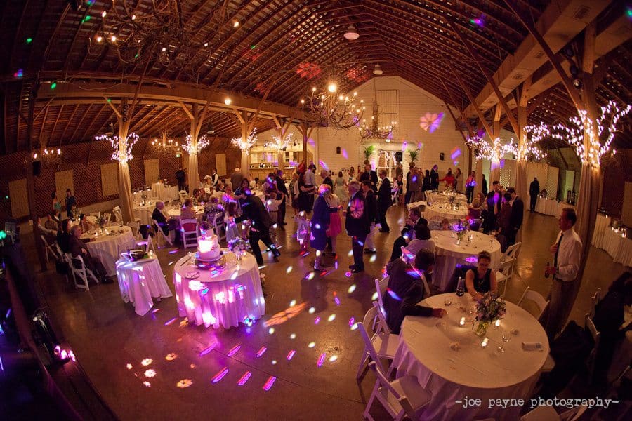 A festive wedding reception in a barn with guests dining and dancing. The warm lighting and colorful disco lights create a lively atmosphere. Several round tables are decorated with white linens and candles, and a decorated cake is on display.