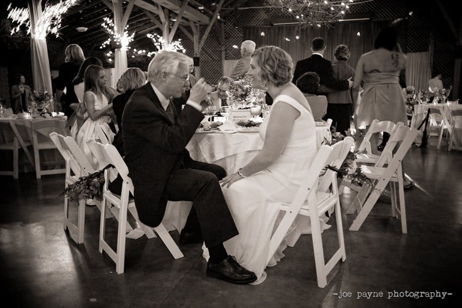 A couple dressed in formal attire sit on white chairs facing each other at a wedding reception. The man is sipping from a glass, while the woman watches. Guests mingle in the background under string lights. The setting is warm and elegant.