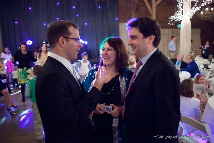 Three people are smiling and conversing at a lively indoor event. A man on the left is gesturing while a woman and another man listen attentively. The background is filled with guests, twinkling lights, and a dark curtain.