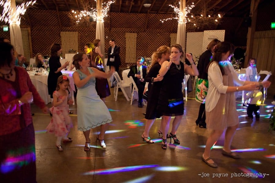 A group of people dance joyfully on a dance floor at a wedding reception. Sparkling lights illuminate the room, and tables with seated guests are visible in the background.