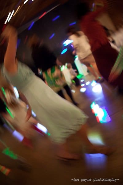 Blurred image of people dancing at a party with colorful lights creating a dynamic, swirling motion effect. The scene captures a sense of energy and movement on the dance floor.