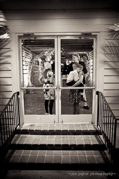 A black and white photo shows a couple embracing and dancing inside a warmly lit building, visible through a glass door. Holiday lights decorate the interior, and two women chat beside the couple. The scene conveys a cozy, festive atmosphere.