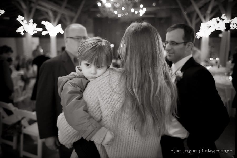A black and white photo of a woman with long hair carrying a young child. The child rests their head on her shoulder, appearing thoughtful. Two men, one in glasses and a suit, stand nearby. The background shows a softly lit indoor venue.