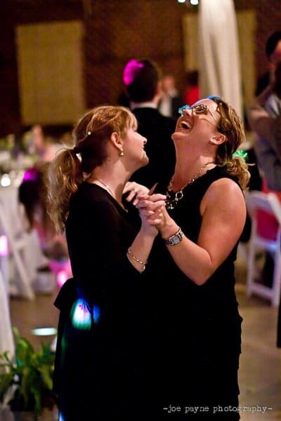 Two women laughing and holding hands while dancing in a dimly lit room. They appear to be enjoying themselves at a festive event, with blurred guests and tables in the background.